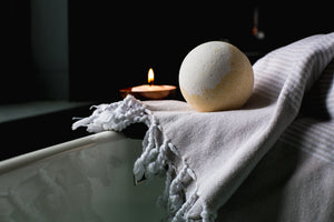 Lights are off in room.  A yellow and white color bath bomb is resting on a white and beige towel on the edge of what could be a bath tub.  A small votive lit candle in a brass color container is to the left on a ledge about 5 feet away.